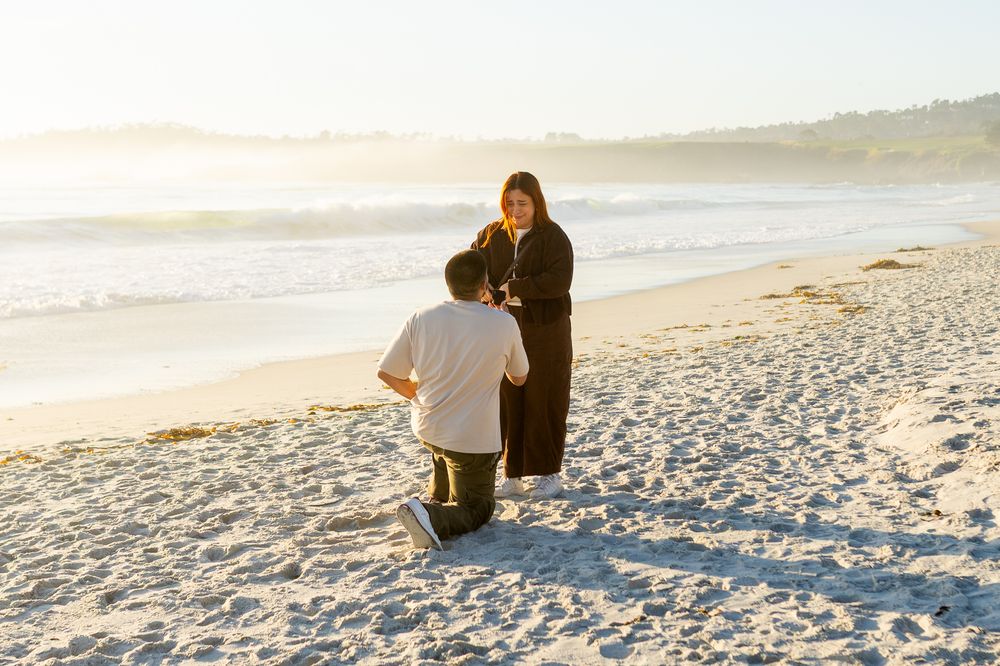 Man kneeling on sandy beach proposing to woman at sunset with ocean waves in background.