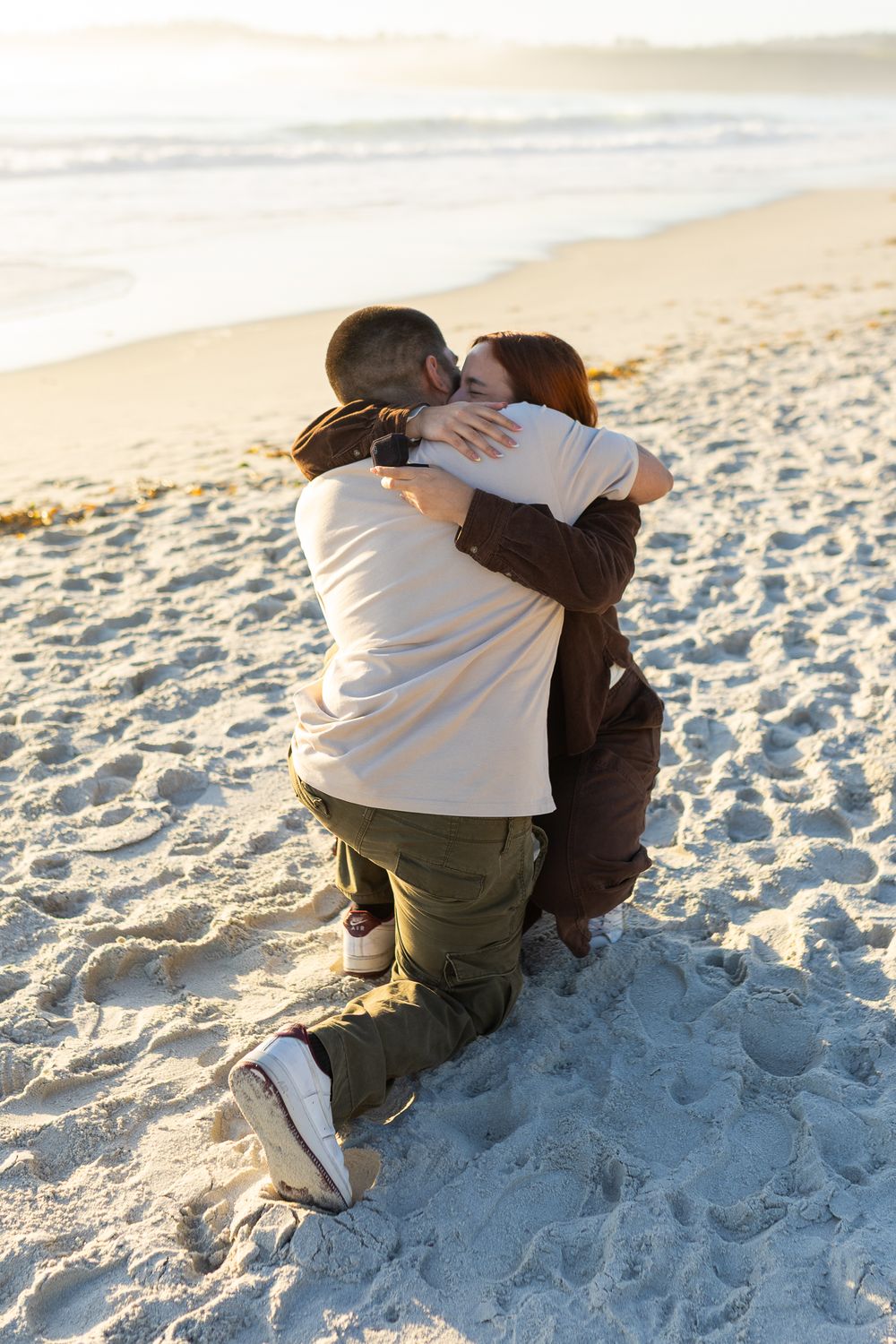 Couple embracing and kissing on a sandy beach at sunset, kneeling together in a romantic moment.