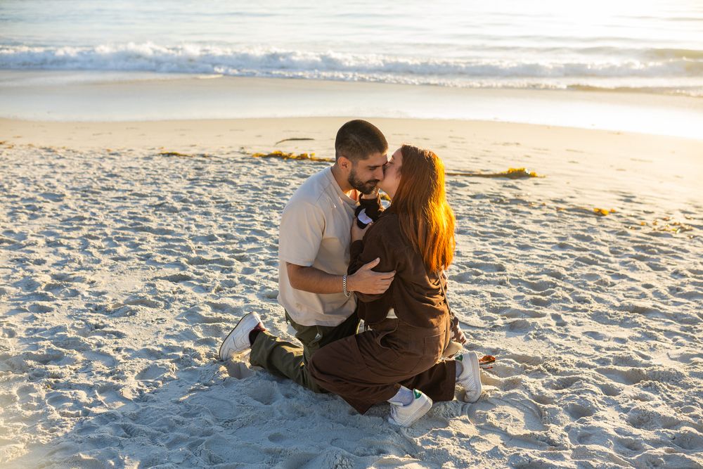 Couple sharing a romantic kiss while sitting on a sandy beach at golden hour with ocean waves in the background.