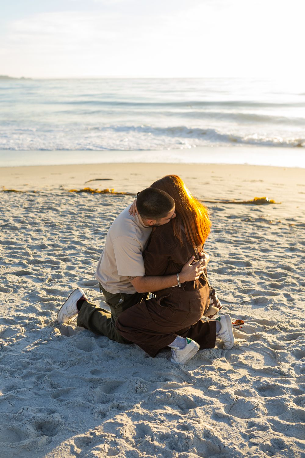 Couple sitting on sandy beach at sunset, sharing a romantic embrace with ocean waves in the background.