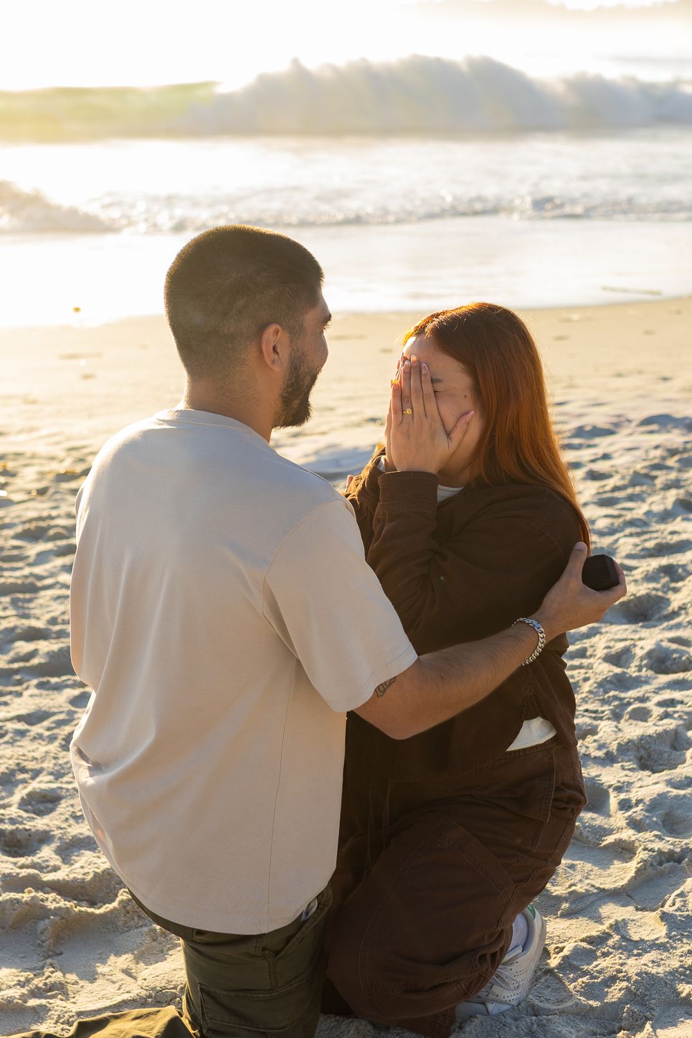 Man proposes to emotional redhead woman on sandy beach at golden sunset, she covers her face in surprise.