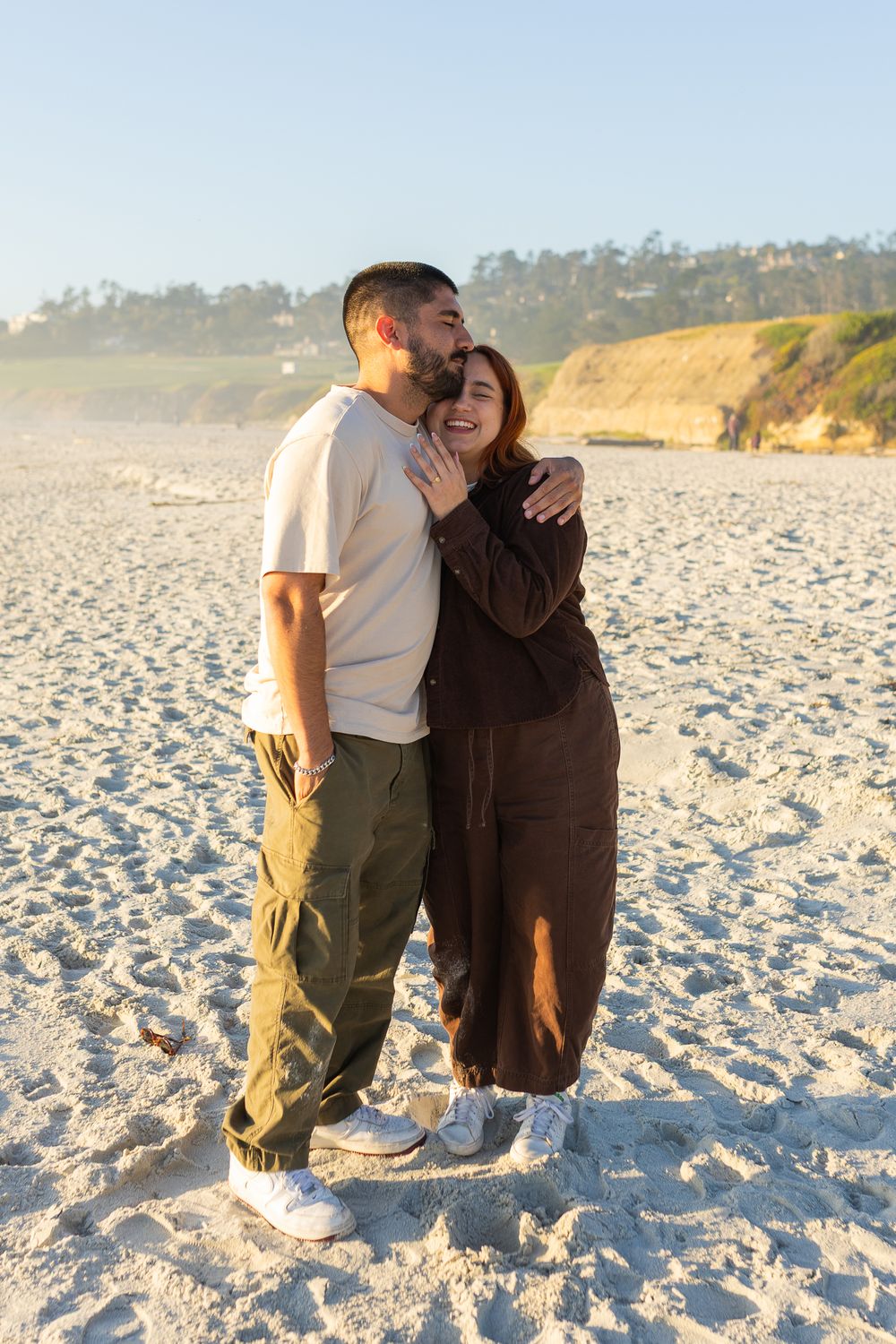 Couple embracing on a sandy beach at golden hour, man kissing woman's cheek as she smiles, with cliffs in background.