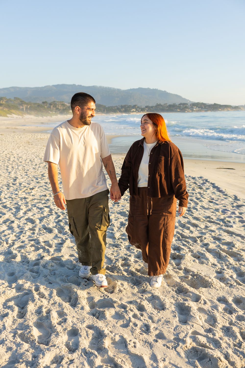 Couple holding hands walking on a sunny beach with ocean waves and mountains in the background.