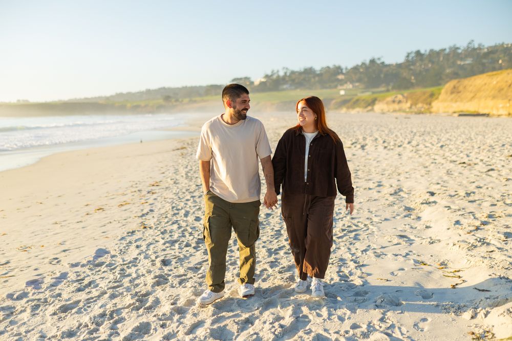 A couple holds hands and walks along a sunny sandy beach, smiling at each other in warm golden light.