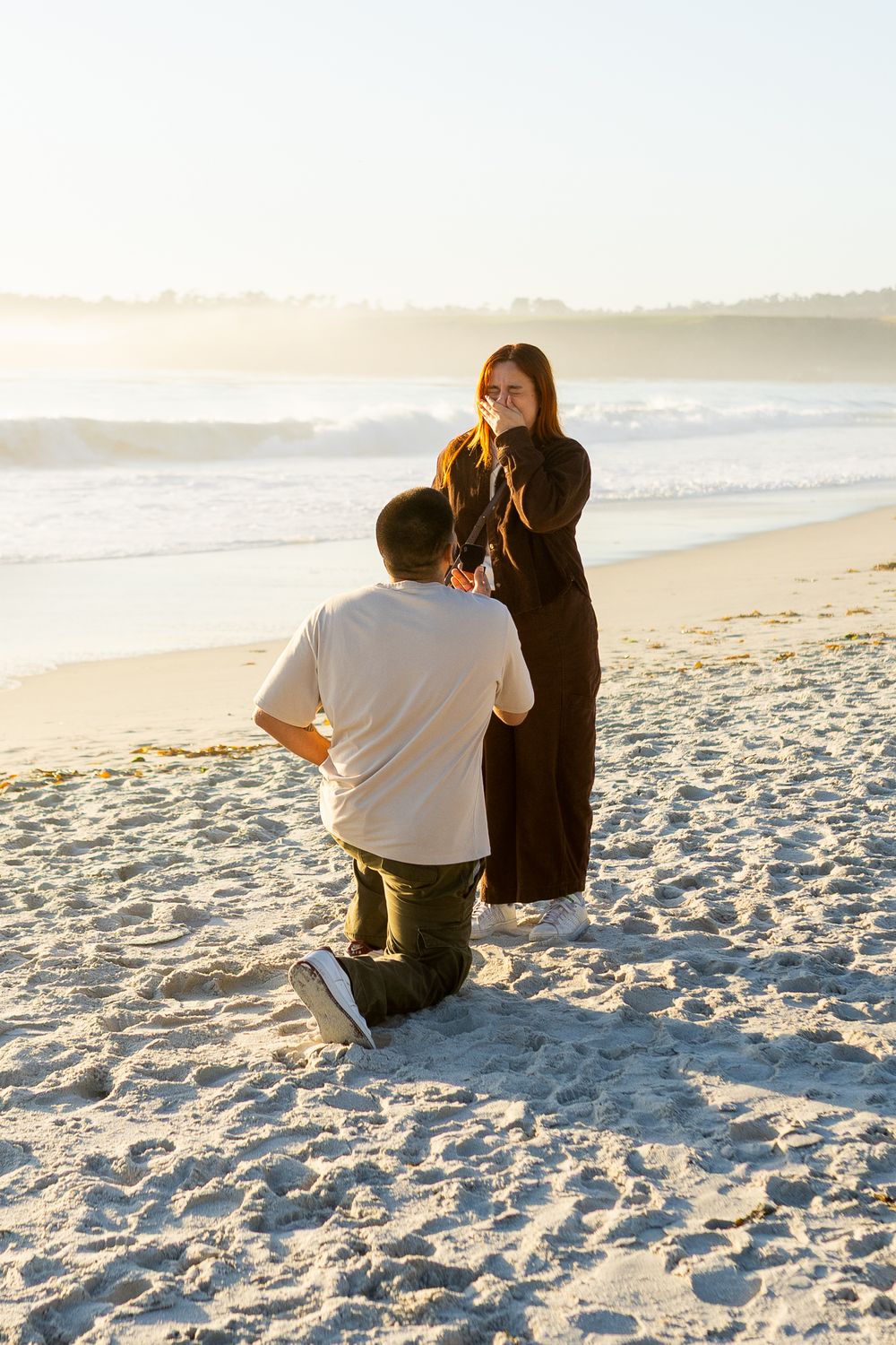 Man proposes on one knee to woman on sandy beach at sunset, as she covers her mouth in surprise.