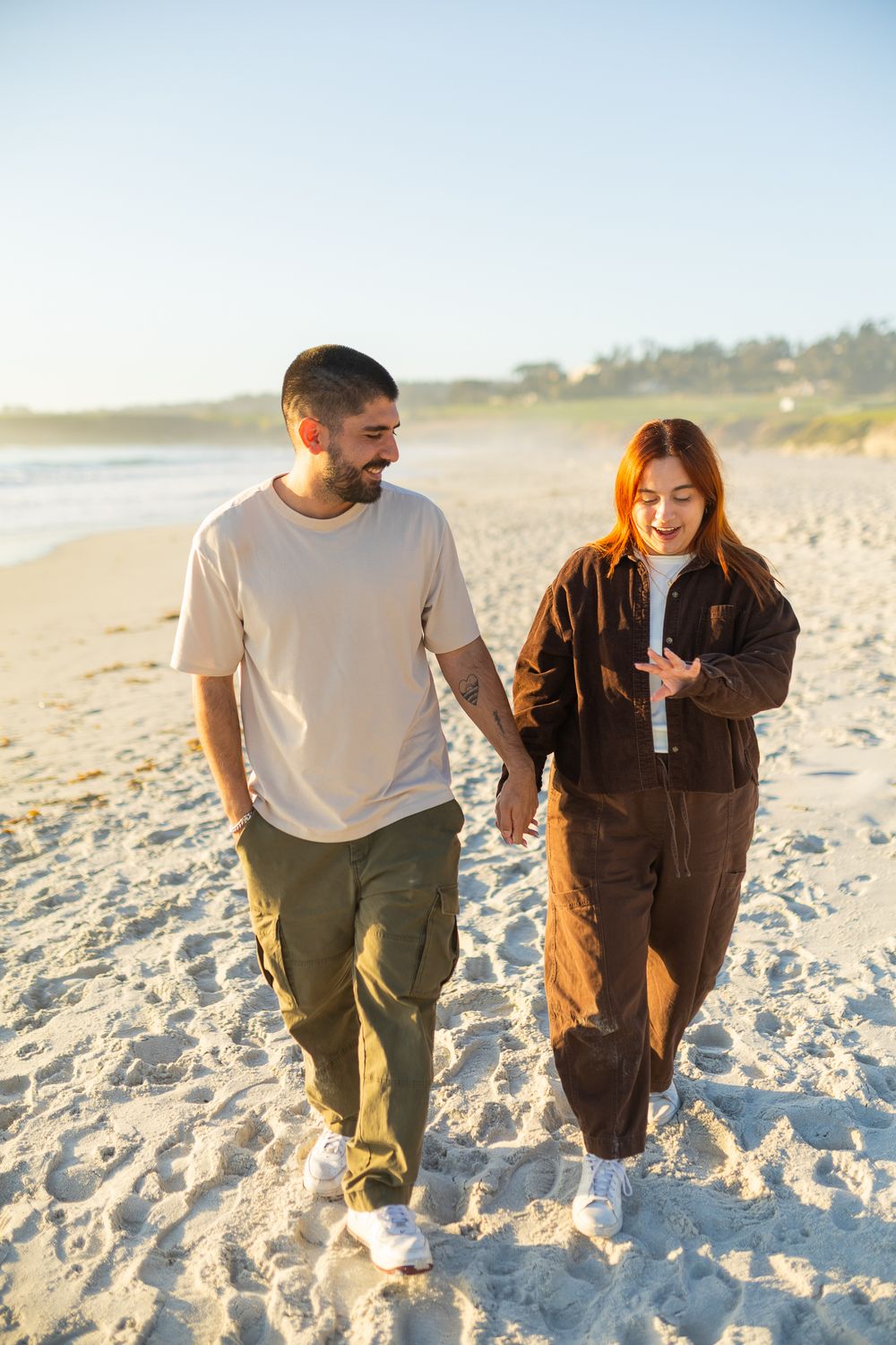 Couple holding hands and walking on a sunny beach, man in beige shirt and green pants, woman in brown jumpsuit.