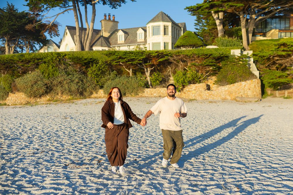 A happy couple holds hands and runs on a sunny white sand beach with a large house and trees in the background.