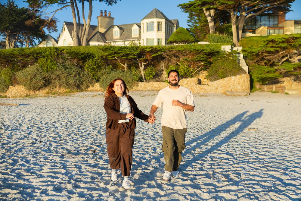 Couple holding hands running on a sunny beach with a large house and cypress trees in the background.