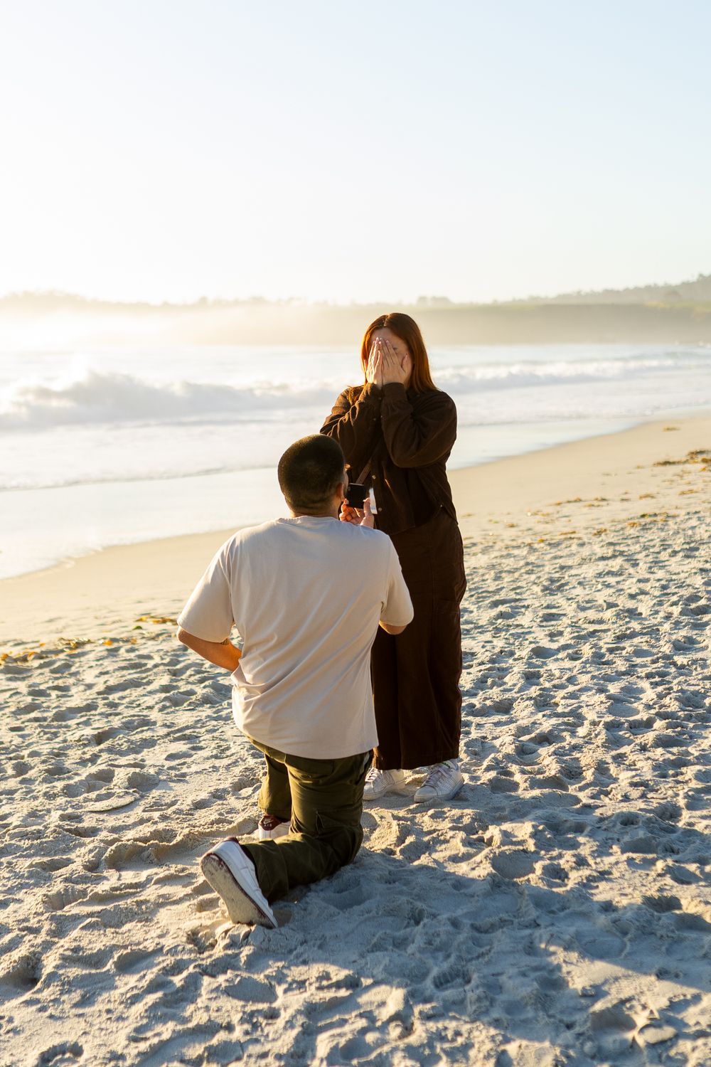 Man kneeling on beach proposing to surprised woman covering her face, golden sunset and ocean waves in background.