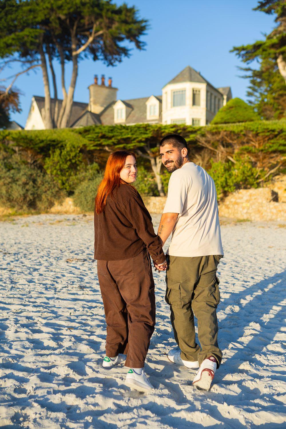 Couple holding hands on sandy beach with luxury home and trees in background during golden hour sunset.