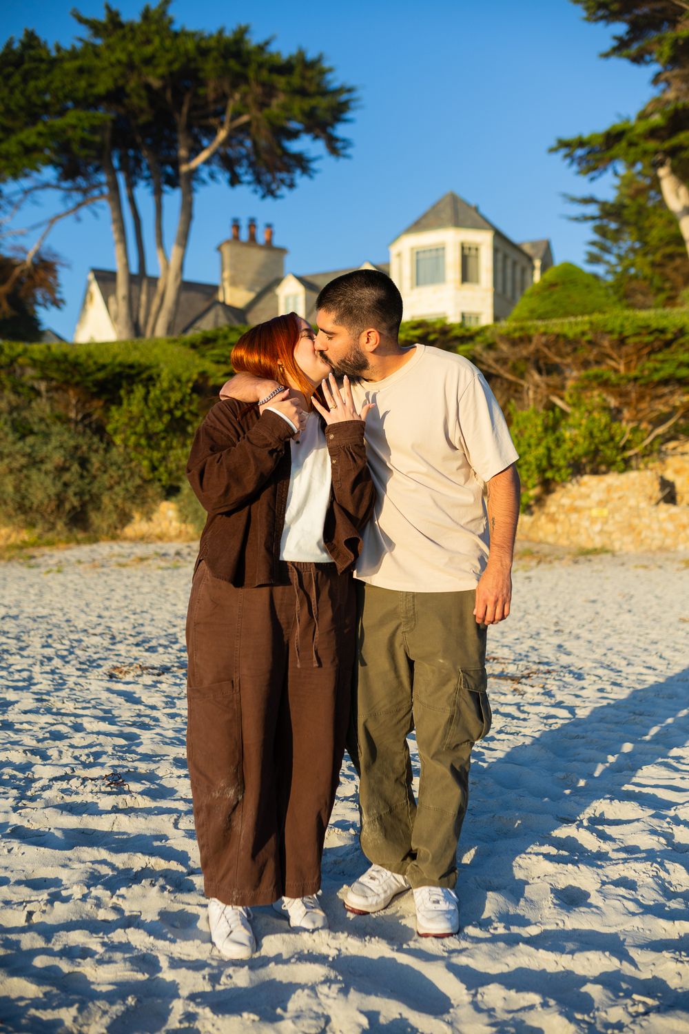 Couple sharing a kiss on a sunny white sand beach with a house and trees in the background.
