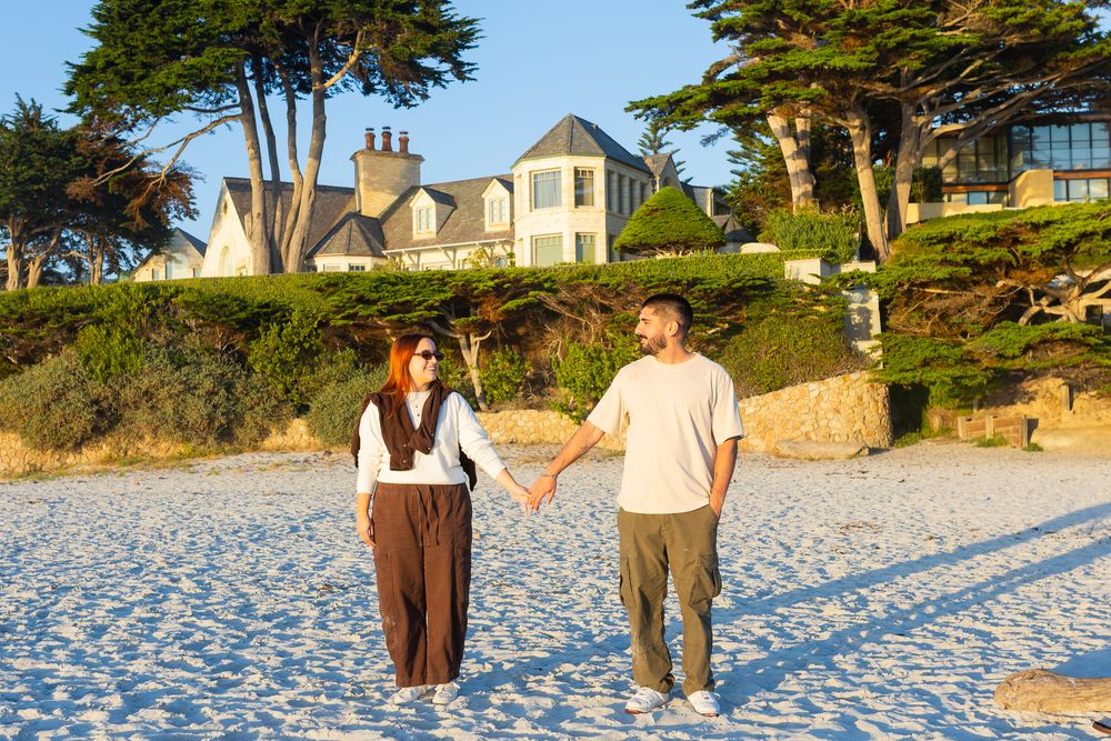 Couple holding hands walking on a sandy beach with a large coastal house and trees behind them.