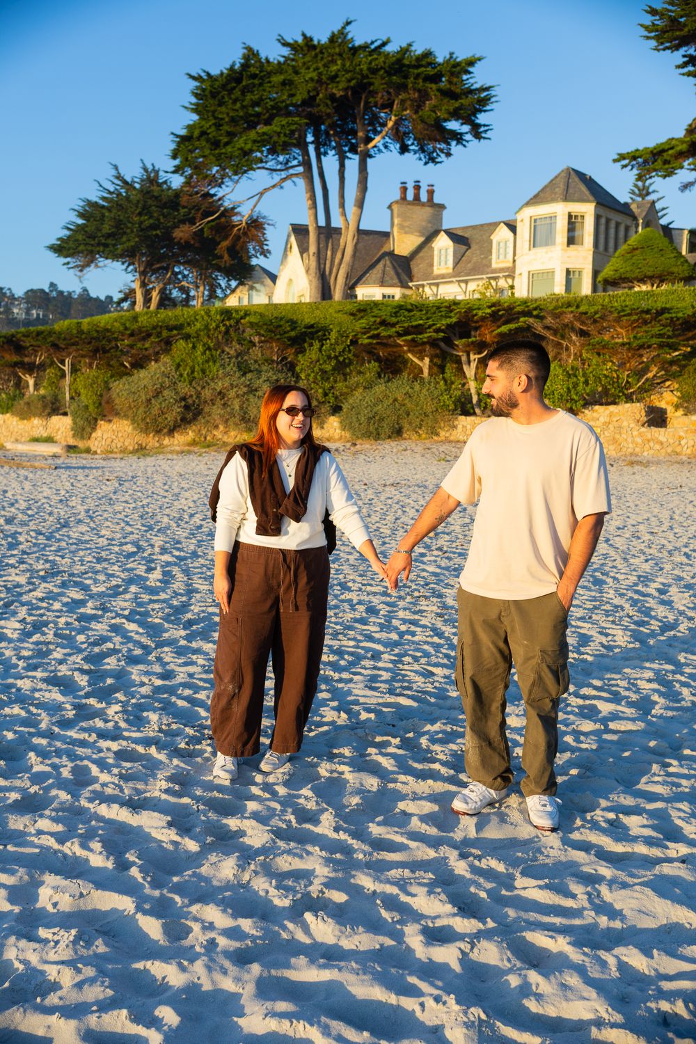 Couple holding hands on a sandy beach with a large coastal house and cypress trees in the background.