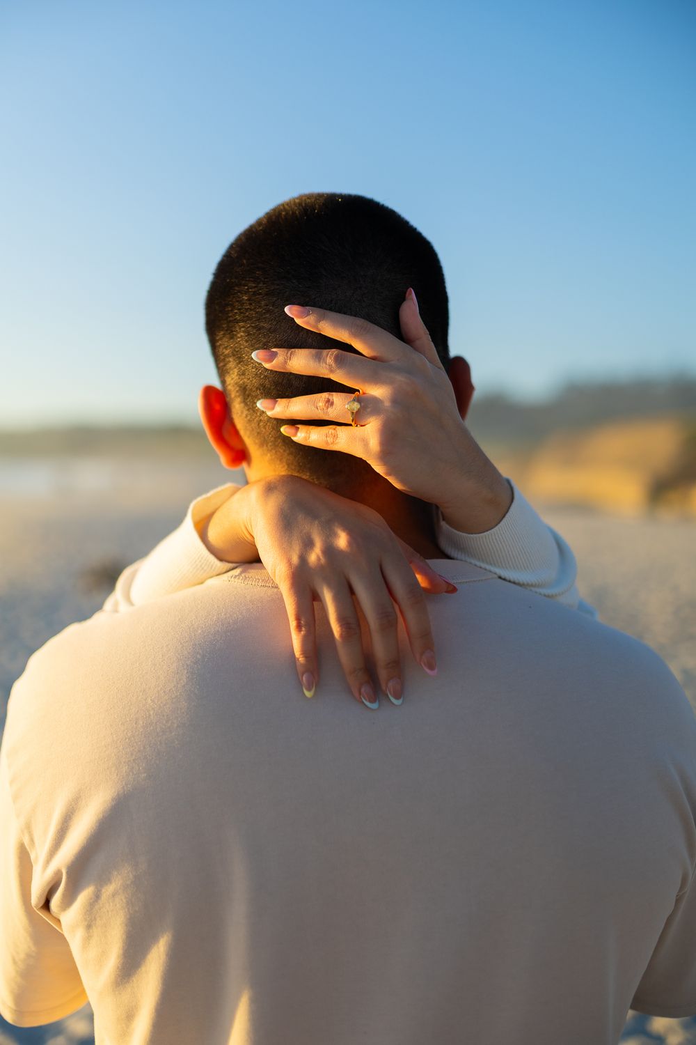 A woman with an engagement ring wraps her arms around a man's neck at sunset on a beach.