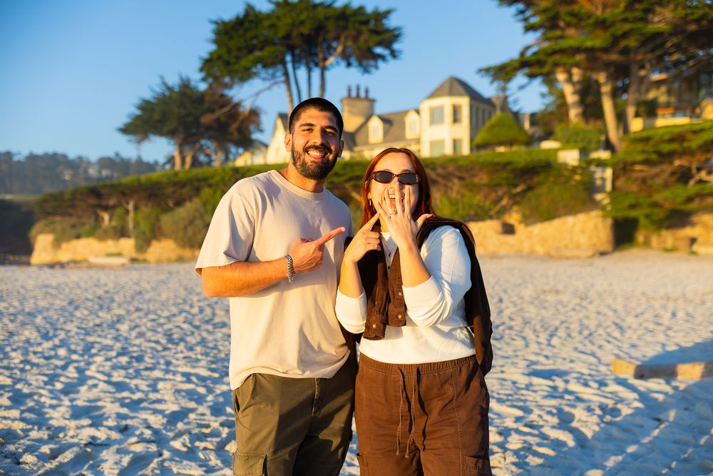 Couple celebrating engagement on sandy beach, woman showing ring, man pointing, with coastal house and trees in background.