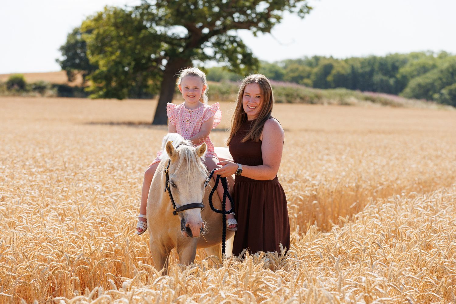 Young girl sitting on a white pony while a woman in a brown dress stands beside her in a golden wheat field on a sunny day.