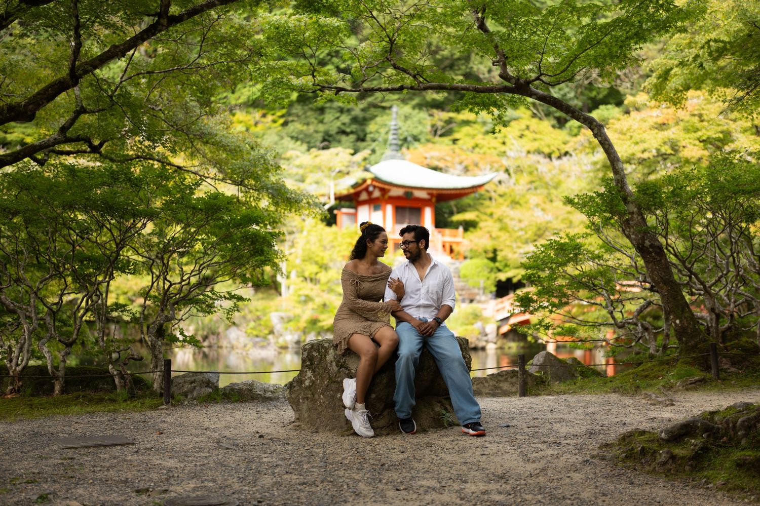 A couple holds hands beside a tranquil pond, gazing at a vermilion Japanese pavilion and arched bridge surrounded by lush green trees.
