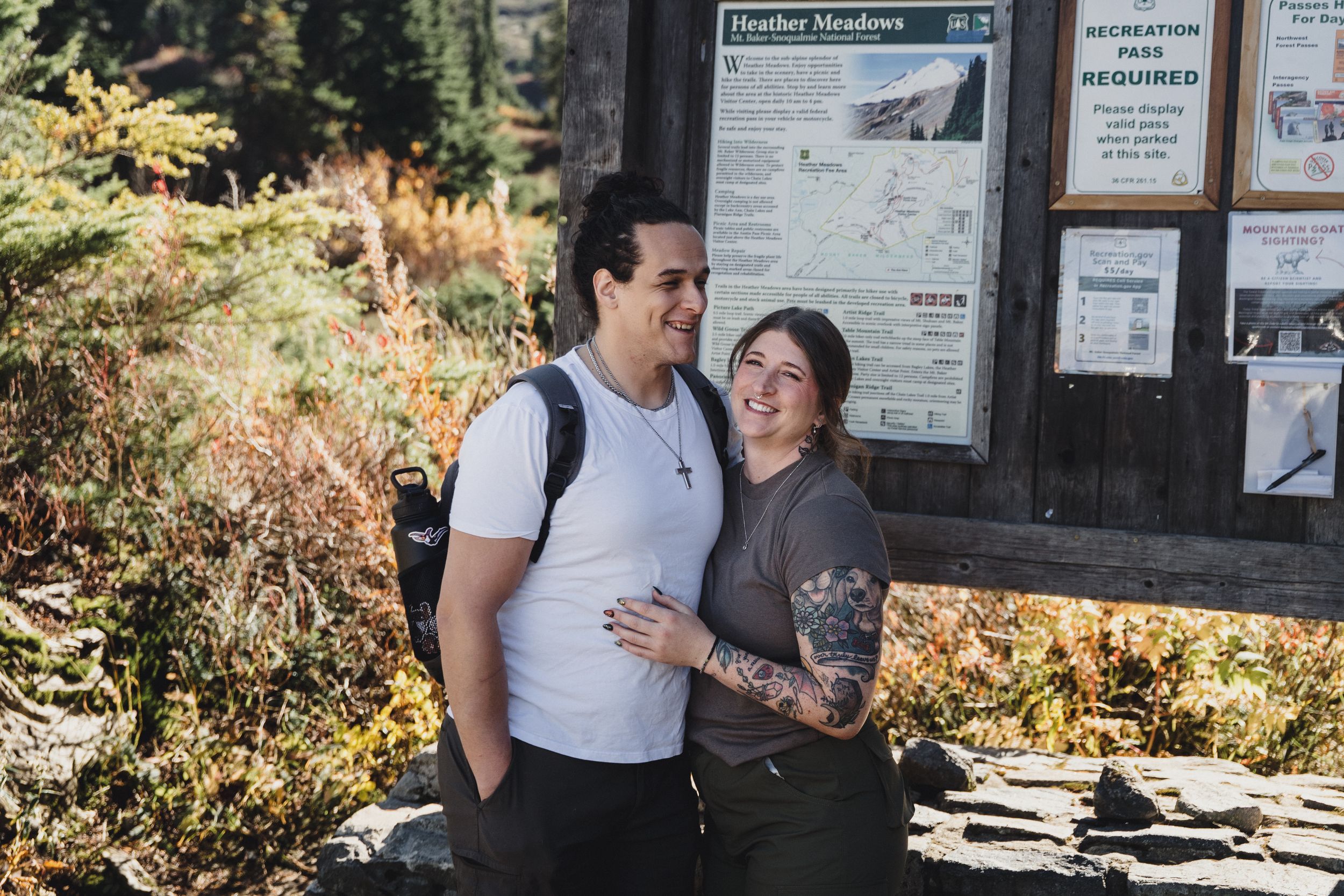 Smiling couple embraces in front of a Heather Meadows trailhead information board surrounded by autumn foliage in the mountains.