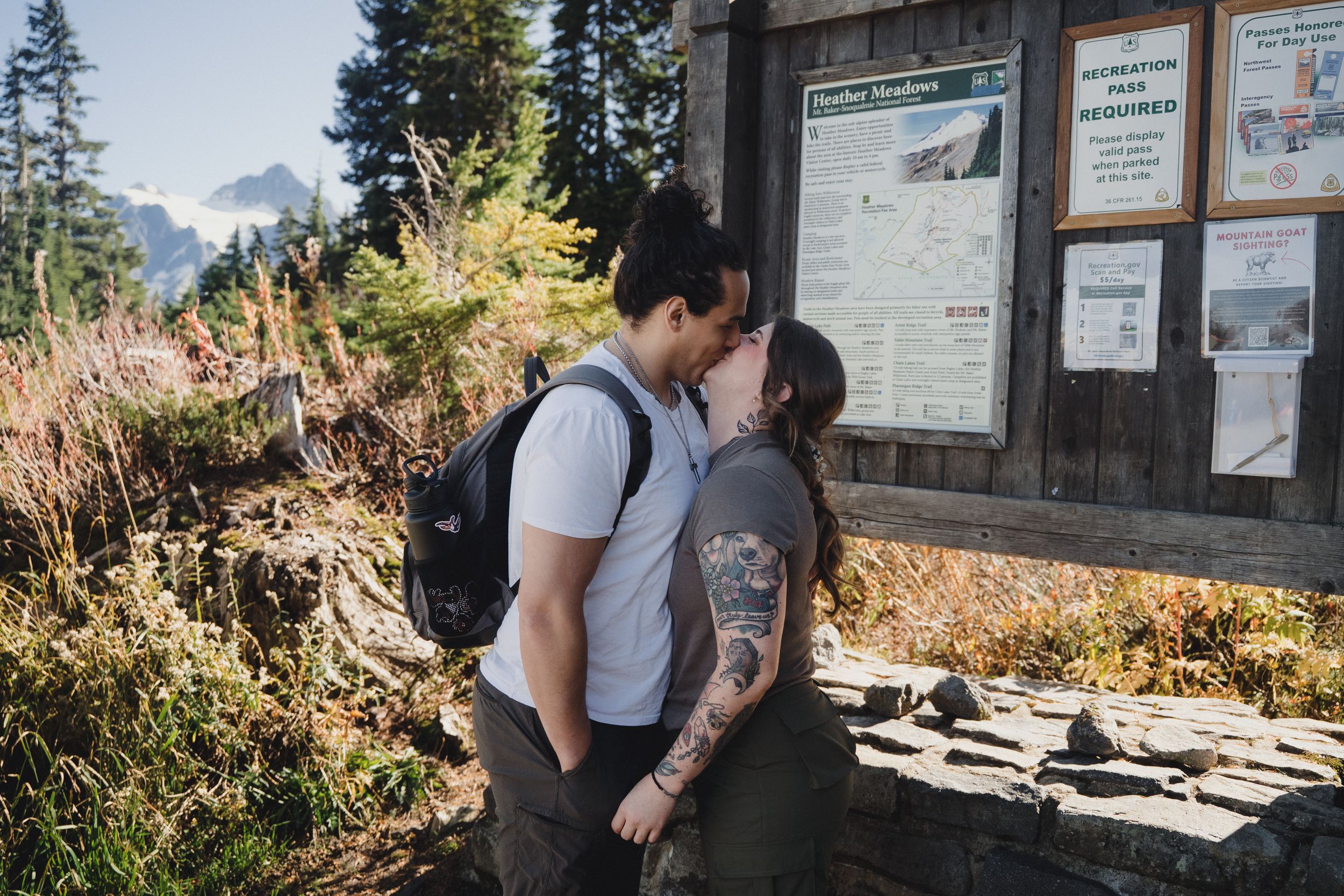 Hiking couple kissing beside a trailhead information board with snowy mountain peaks and evergreen trees in the background.