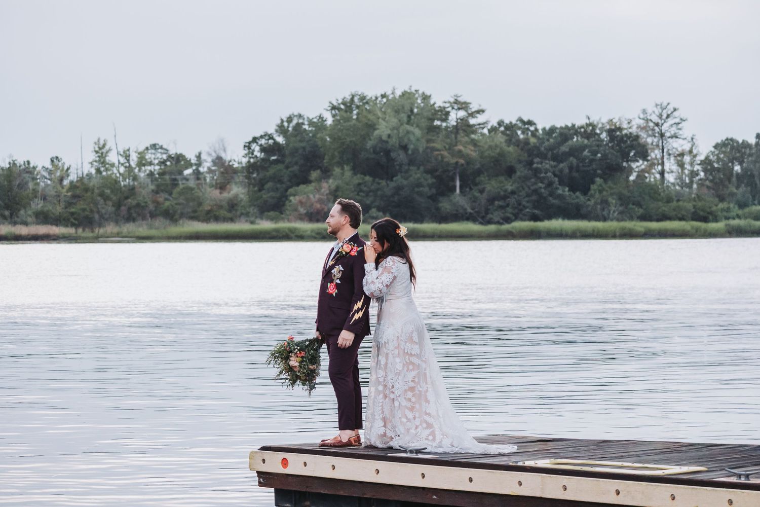 Bride in a lace gown and groom in a dark suit holding a bouquet stand on a wooden dock overlooking a calm lake at dusk.