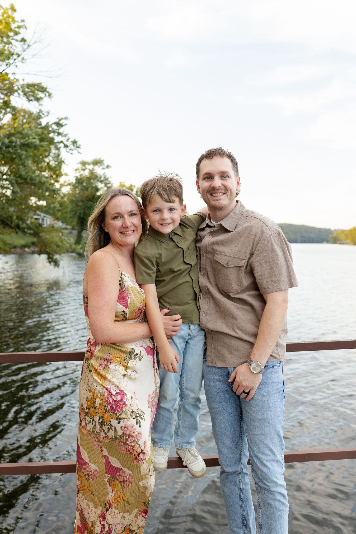 Smiling family of three, a woman in a floral dress, a man, and a young boy, pose together on a lakeside dock at sunset.