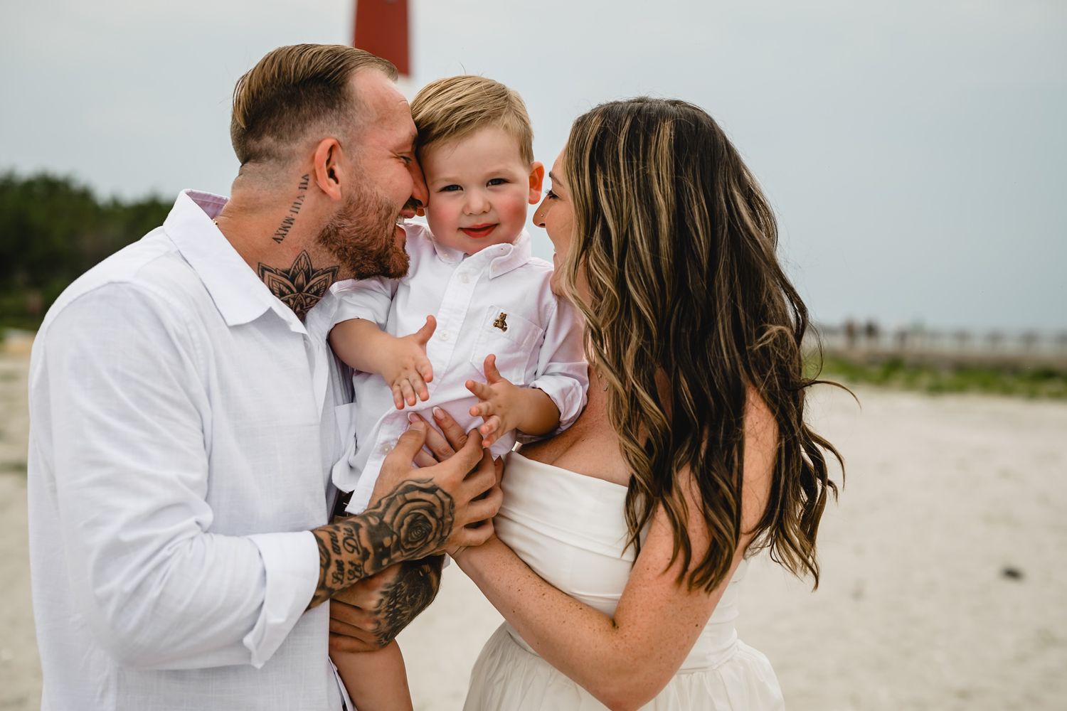 A tattooed father and brunette mother kiss their smiling toddler son on a sandy beach in white outfits.