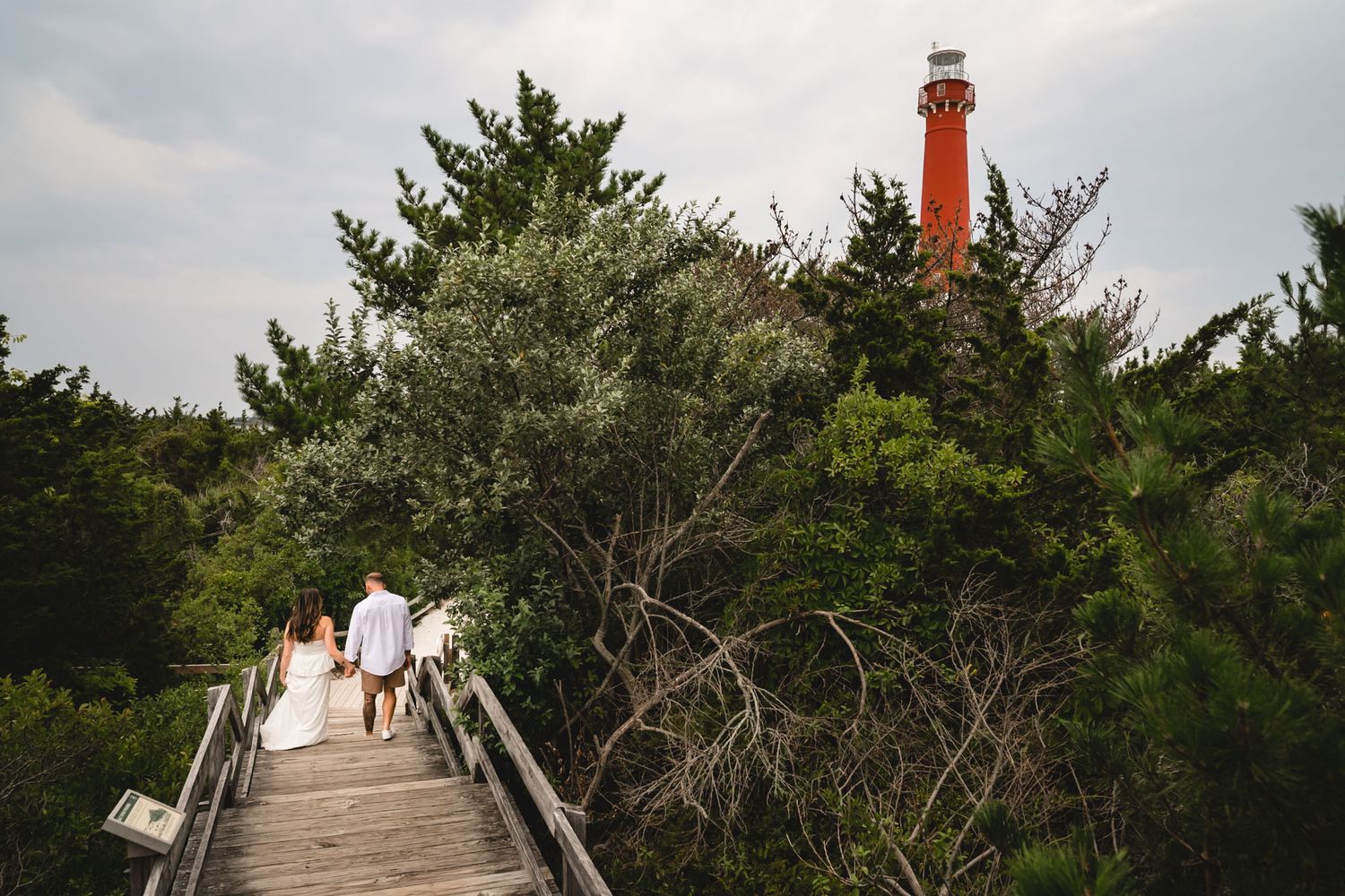 Couple walks along wooden boardwalk path surrounded by lush trees toward a red lighthouse on their wedding day.