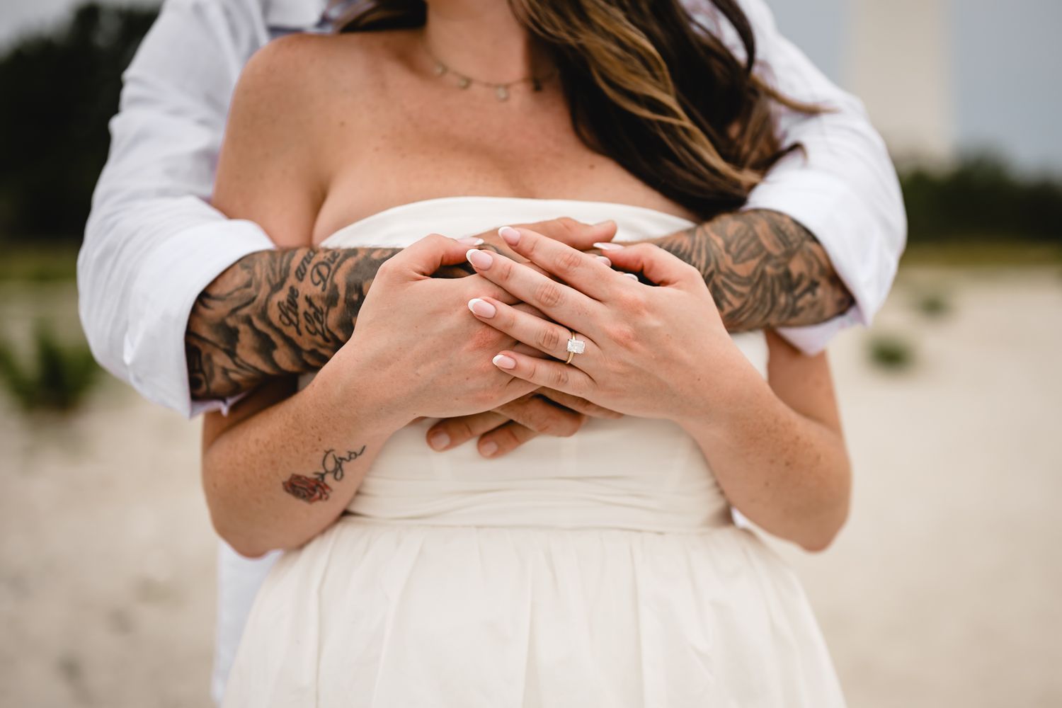 Couple embracing on beach, tattooed arms wrapped around bride in white dress, showing wedding ring.