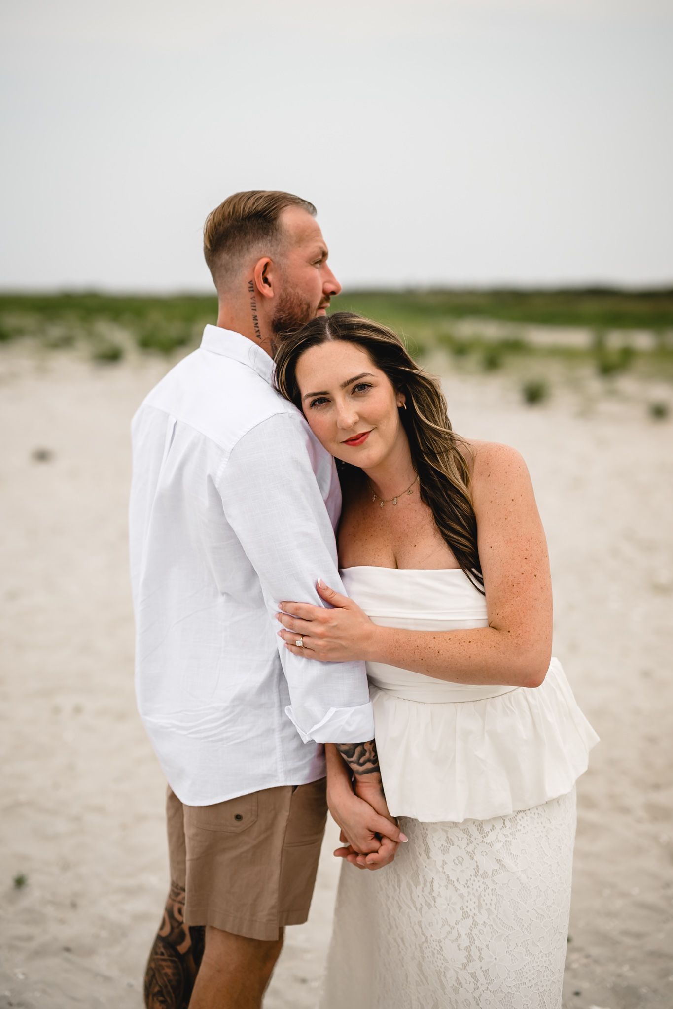 A couple embraces on a sandy beach, the woman in a white lace dress smiling at the camera while the man holds her close.