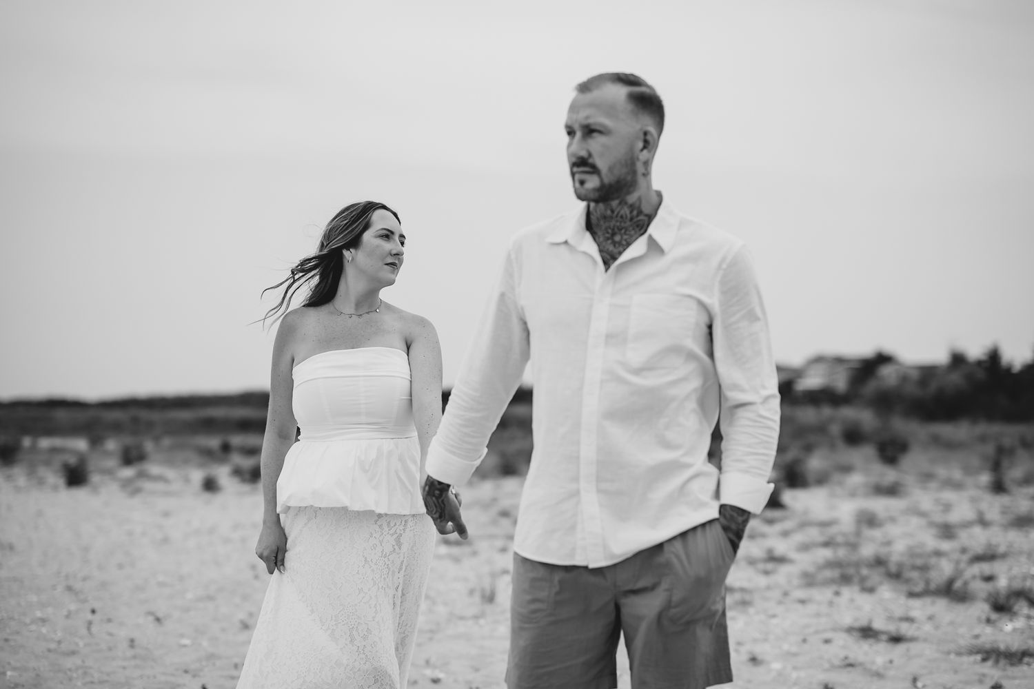 Black and white photo of a couple holding hands on a sandy beach, woman in white dress, tattooed man in white shirt.