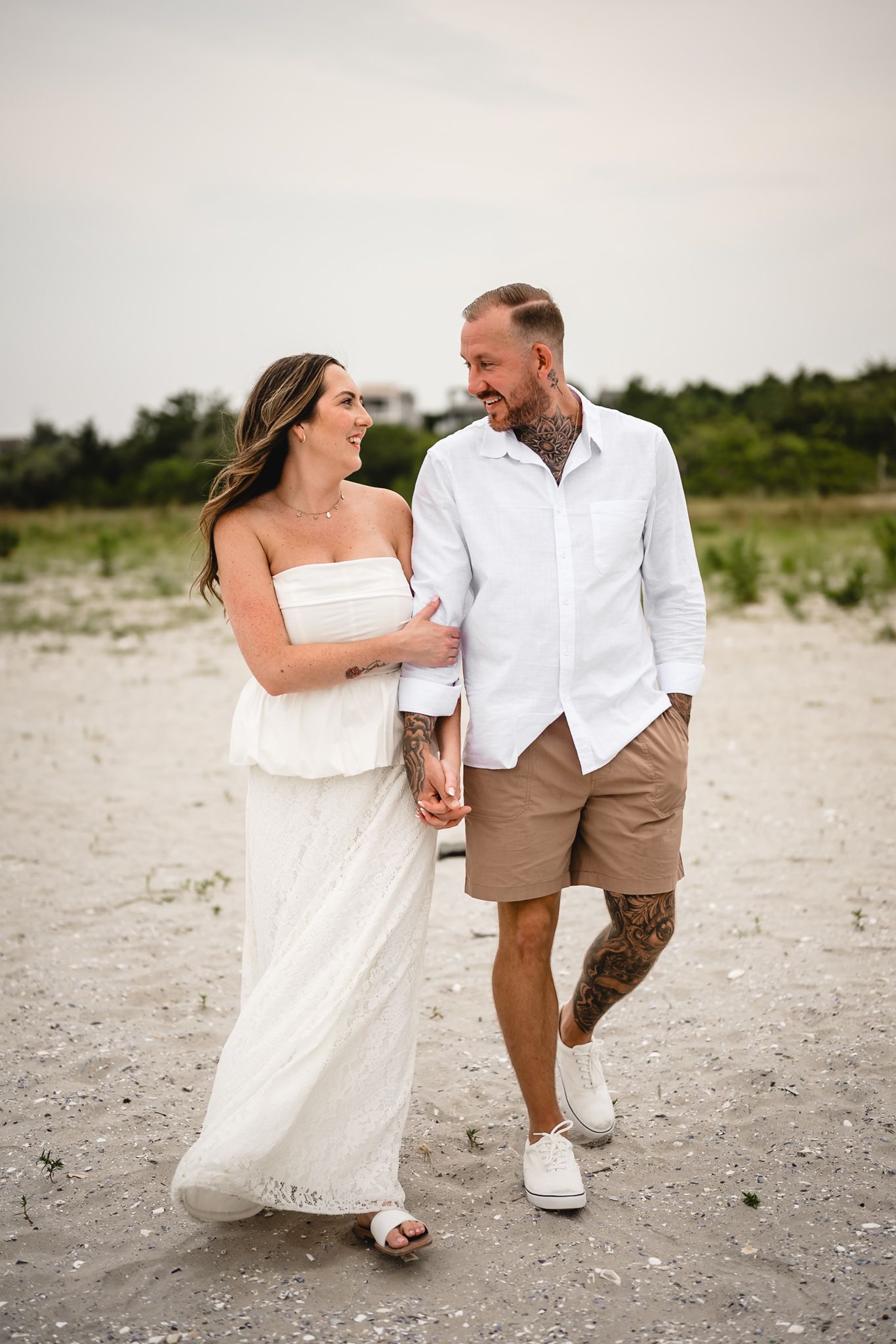 A couple walks hand-in-hand on a sandy beach, smiling at each other, dressed in white and tan casual summer outfits.