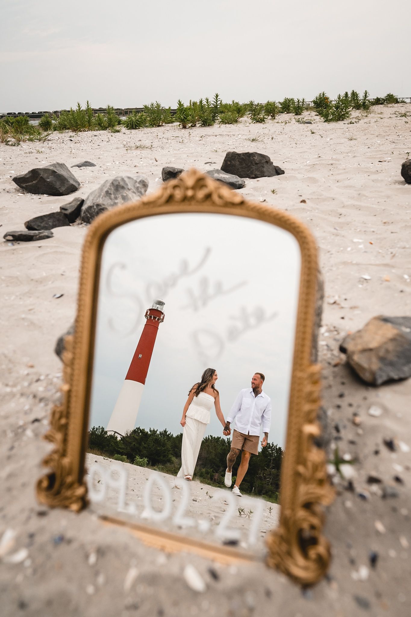 A golden ornate mirror on a sandy beach reflects a couple holding hands near a lighthouse with Save the Date written on it.