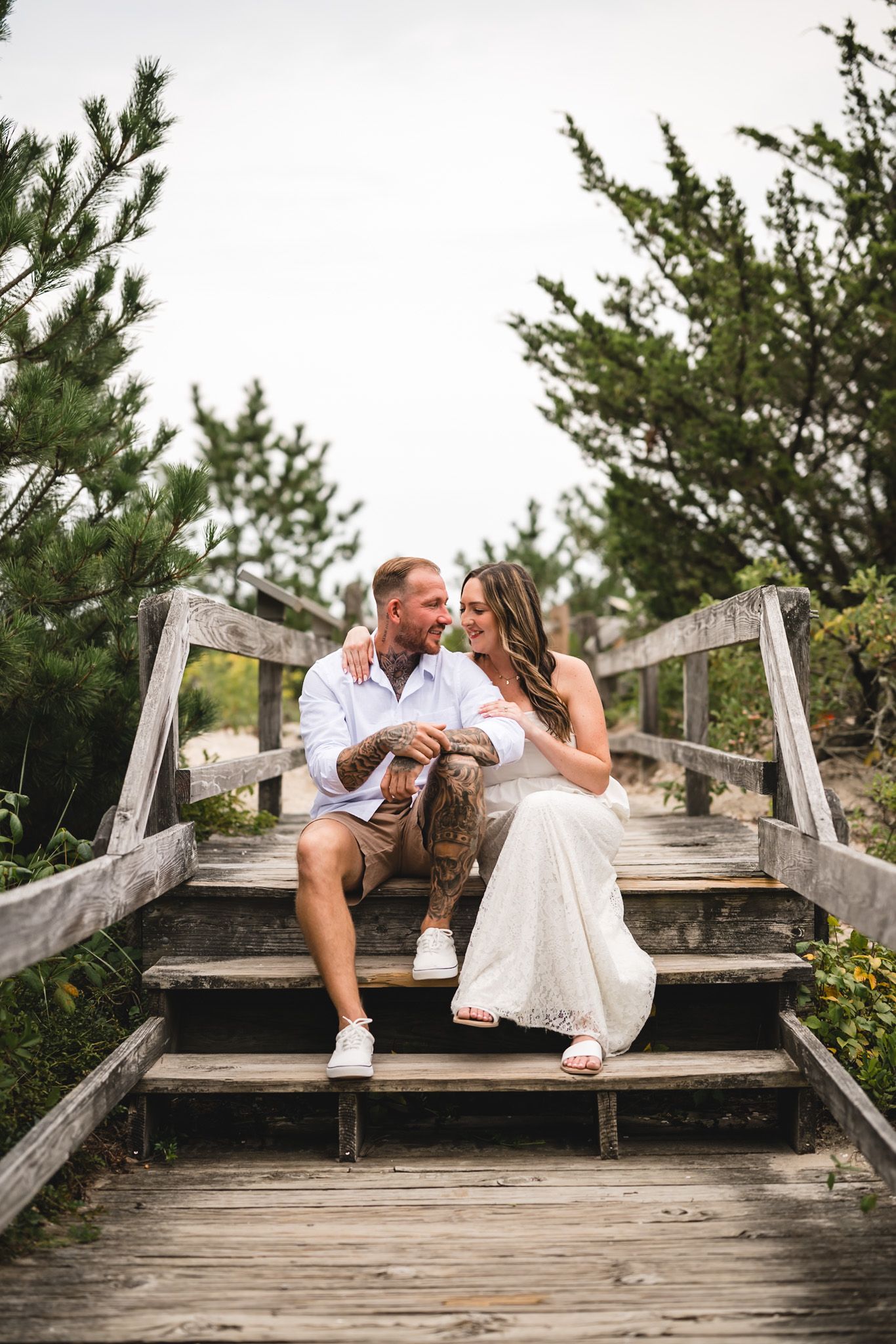 A couple sits together on wooden boardwalk steps, smiling at each other, surrounded by lush green trees.