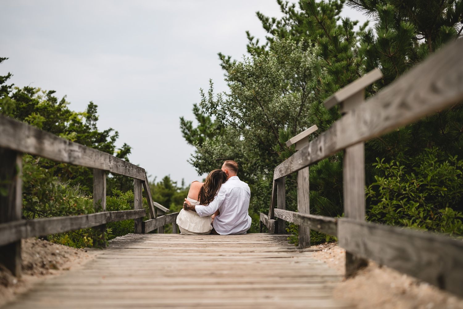 A couple sits together on a wooden boardwalk path surrounded by lush green trees at a serene beach setting.