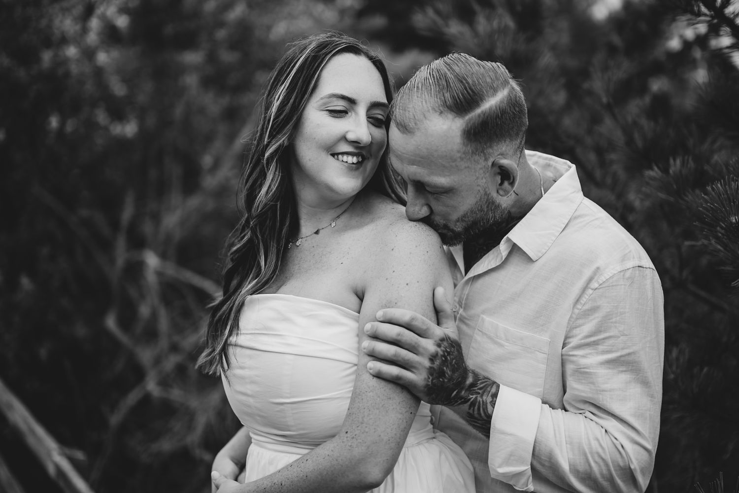 Black and white photo of a couple embracing outdoors, man kissing woman's shoulder, she smiles softly in a strapless dress.