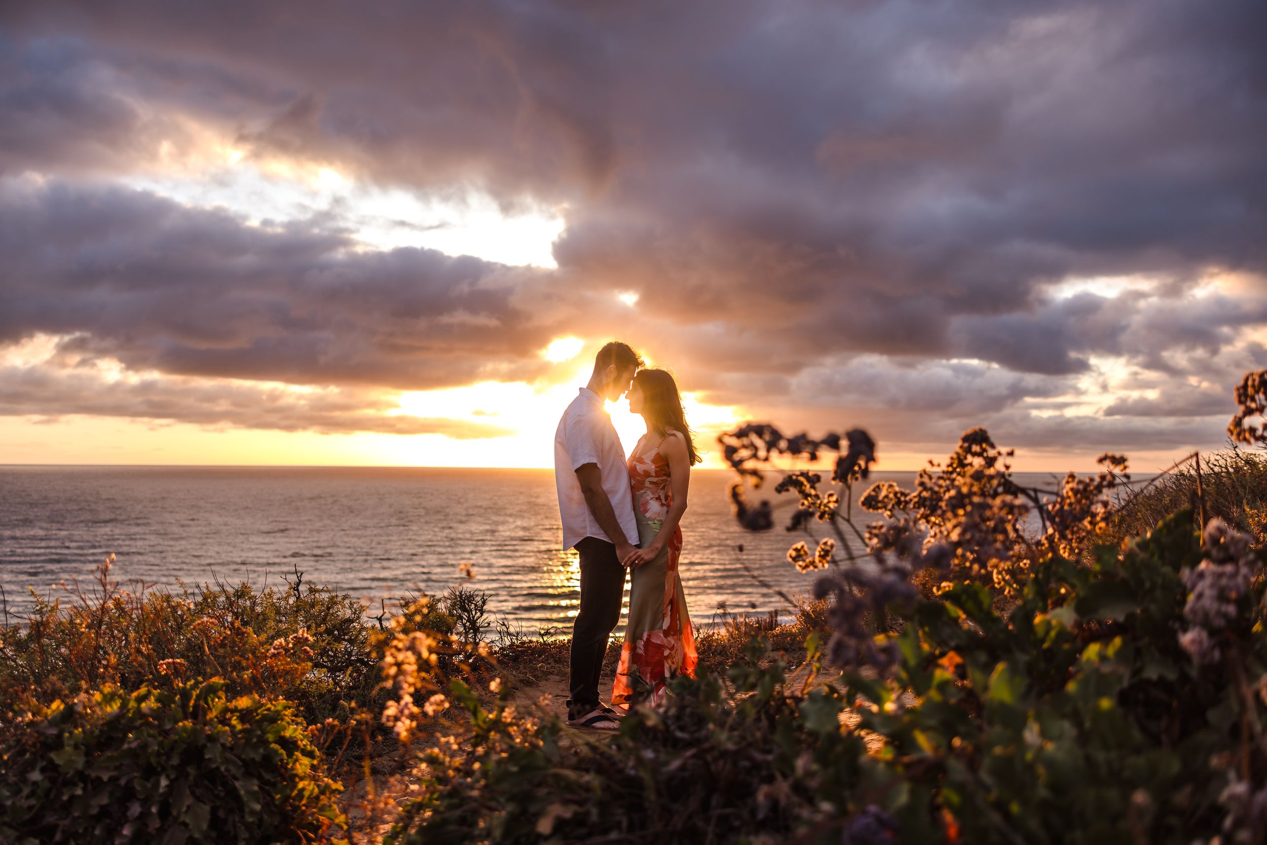 Couple sharing a romantic moment at sunset by the ocean, surrounded by wildflowers under a dramatic cloudy sky.