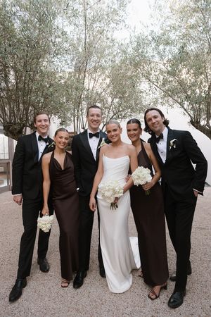 Wedding party poses outdoors in elegant attire, bride in white gown holding white bouquet, bridesmaids in brown dresses.