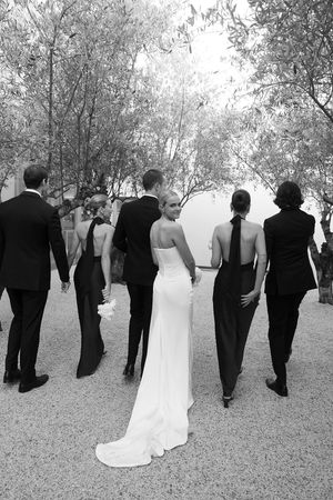 Black and white wedding party walking through tree-lined path, bride in white gown with bridesmaids in black dresses.