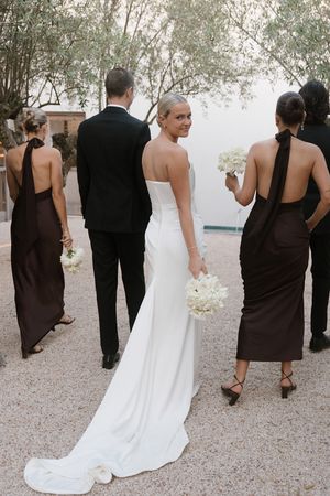 Bride in strapless white gown turns to smile, flanked by bridesmaids in brown halter dresses and groom in black tuxedo.