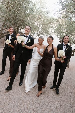 Wedding party walks outdoors, bride in white gown with groomsmen in black tuxedos and bridesmaid in brown halter dress.