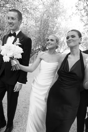 Black and white photo of smiling bride in white strapless gown walking with groom in tuxedo and bridesmaid in halter dress.