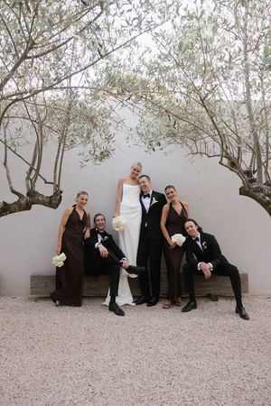 Wedding party poses playfully outdoors with bride in white gown, groom in tuxedo, bridesmaids in brown dresses, groomsmen in dark suits.