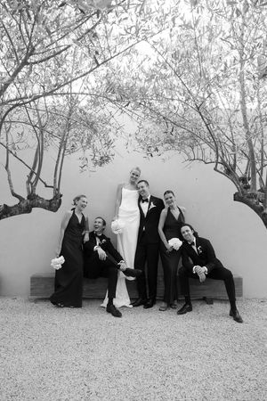 Black and white photo of wedding party posing playfully on bench under trees, bride in white gown with bridal party.