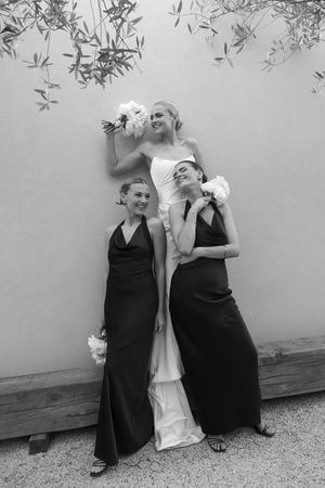 Black and white photo of bride in white gown with two bridesmaids in black dresses holding white bouquets against a wall.