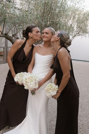 Bride in white gown receives kisses from two bridesmaids in dark brown dresses, all holding white floral bouquets.