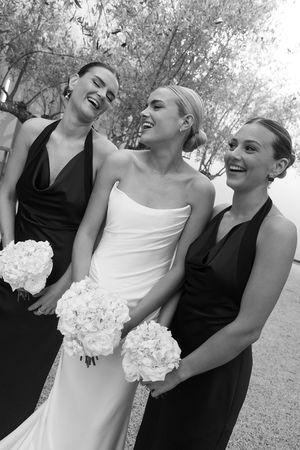 Black and white photo of a bride in white gown laughing with two bridesmaids in black dresses holding white floral bouquets.
