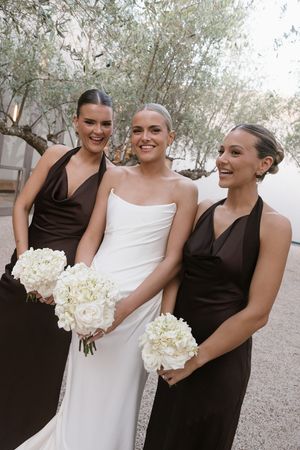 Bride in white gown with two bridesmaids in brown dresses holding white floral bouquets outdoors.