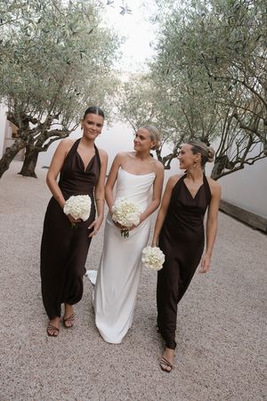 Bride in white gown with two bridesmaids in dark brown halter dresses holding white bouquets outdoors.
