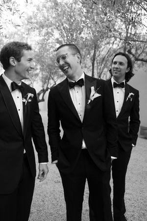 Three groomsmen in black tuxedos with boutonnieres laugh together outdoors in a black and white wedding photo.