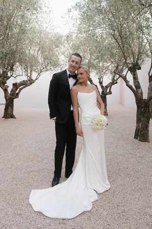 A bride and groom pose together among olive trees, she in a white gown holding flowers, he in a black tuxedo.